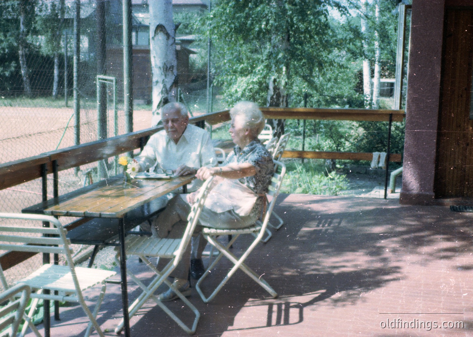 Two elderly individuals seated at an outdoor café table, likely mid-20th century (). Man in light-colored shirt, woman in patterned dress. Foldable metal chairs and wooden picnic-style table. Lush greenery and trees in background, suggesting a park or garden setting. Glass partition with metal railings. é
