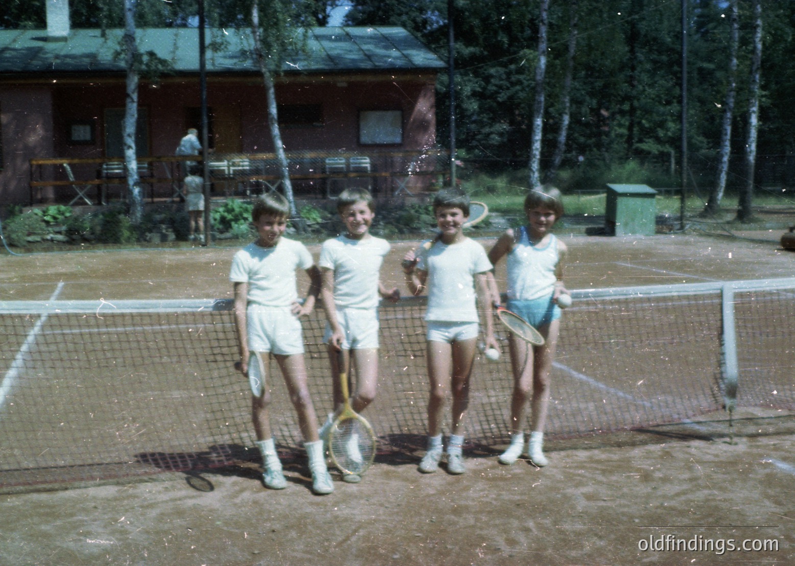 Four young boys in 1970s-style tennis attire pose on a clay court, holding vintage rackets. Wooden cabins and birch trees frame the scene, suggesting a rural or resort setting. Light rain creates a misty effect.