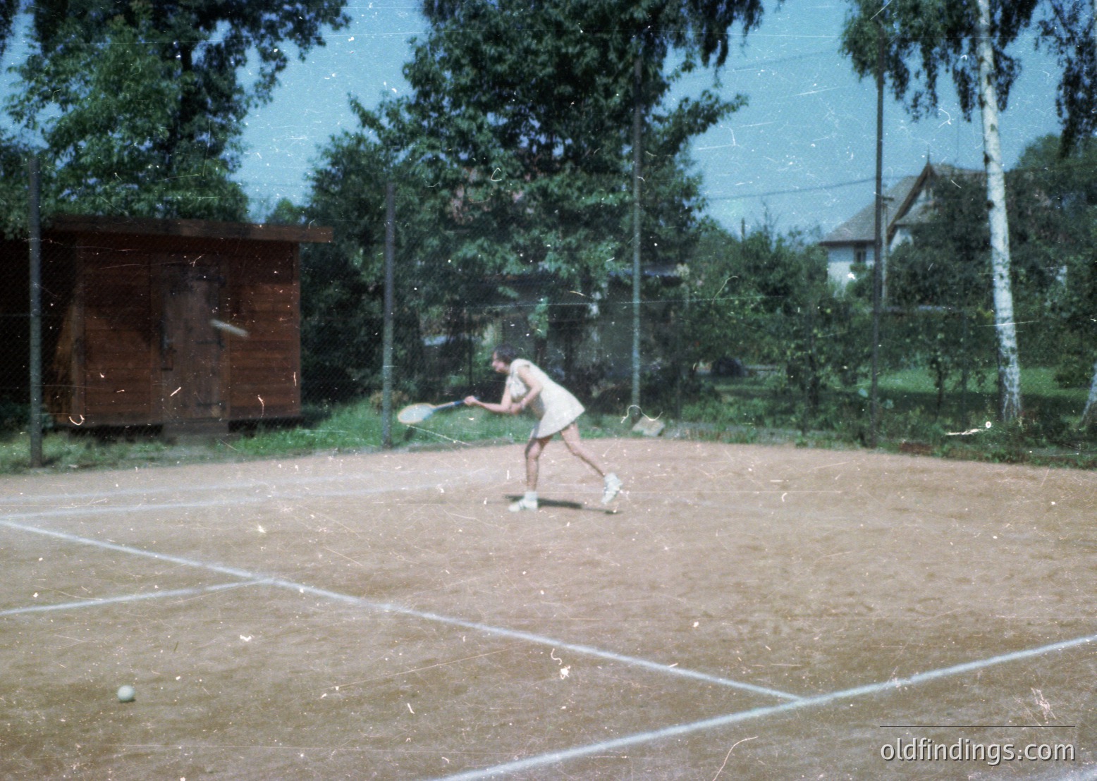 Vintage tennis court scene with player mid-swing, likely 1960s-70s. Clay surface, wooden changing hut, and lush greenery in background. Casual, outdoor recreational setting.