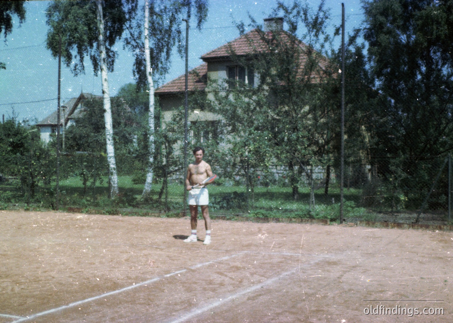 Vintage tennis court in a residential area, featuring a single male player in 1970s-style athletic wear (white shorts, tank top, socks, and sneakers). The court’s clay surface shows visible wear, and a two-story house with a tiled roof and birch trees frames the background. Likely Eastern European setting.