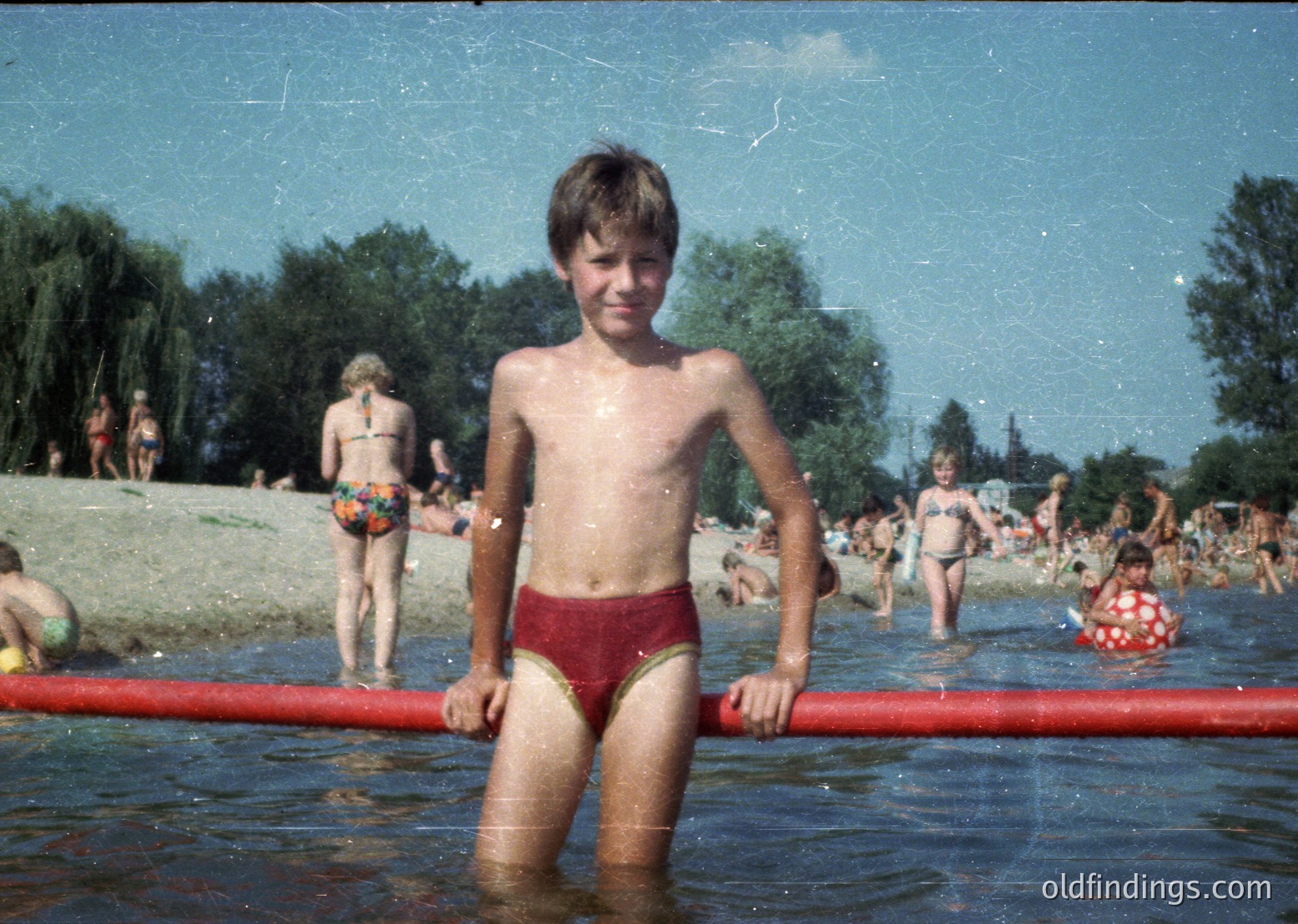 Vintage 1970s beach scene with boy in red swim trunks posing at poolside barrier. Crowded public pool with lush green trees in background. Candid, nostalgic summer vibes.