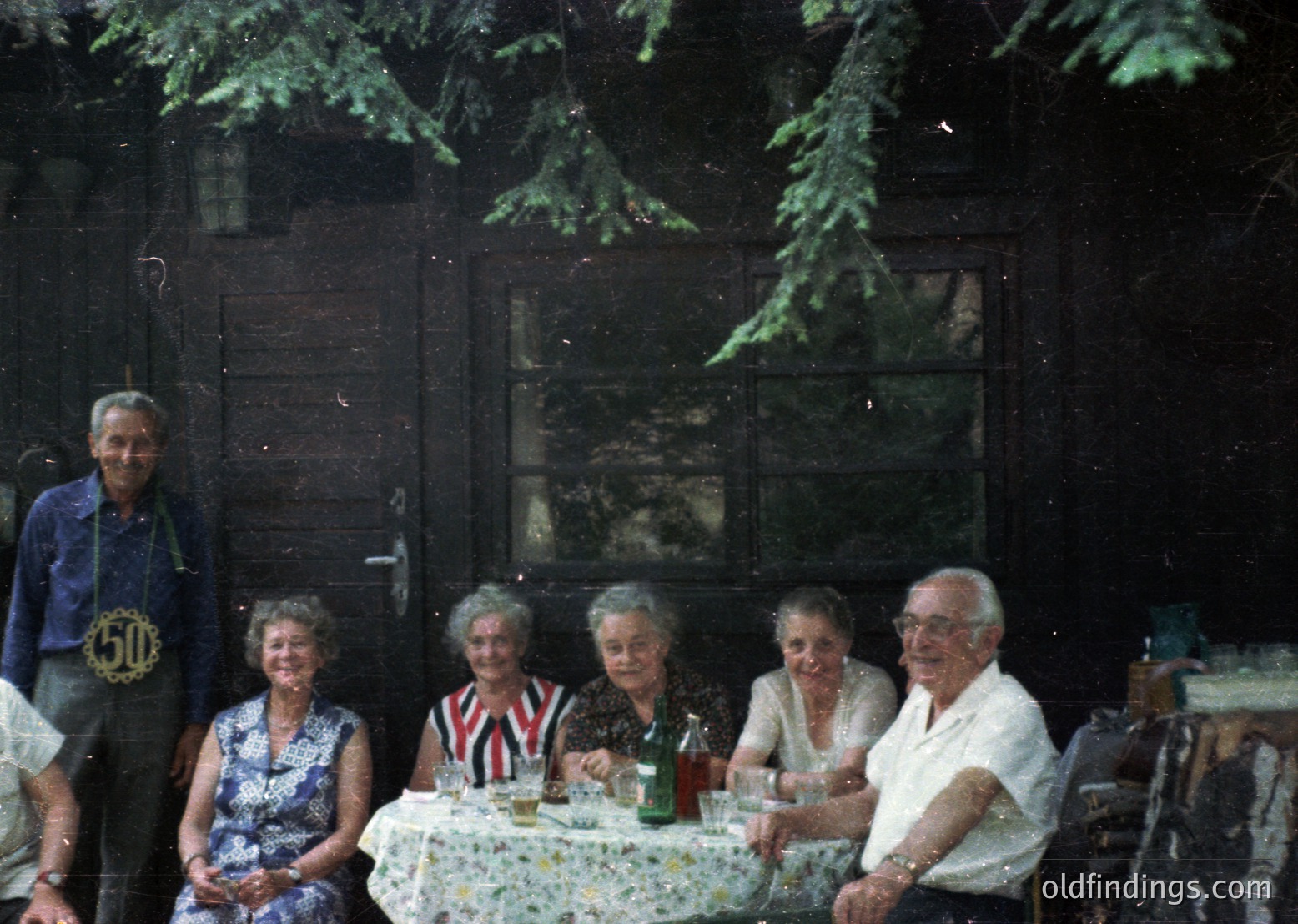 Vintage group portrait in rustic wooden cabin, likely 1970s. Six adults seated at a table with floral tablecloth, posing for a celebratory occasion. Men wear button-down shirts; women in patterned dresses. One man wears a "50" lapel pin. Green glass bottles and a pitcher suggest a social gathering. Moss-covered windows and wooden beams add to the cabin’s charm.