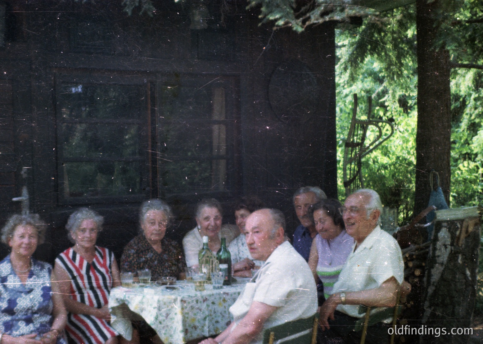 Vintage group portrait of nine elderly individuals seated at a rustic wooden table in a shaded outdoor setting, likely a garden or courtyard. The scene features floral-patterned tablecloths, vintage clothing (1960s–1970s), and a bottle of wine. Wooden chairs and a wooden structure with glass panes frame the composition.