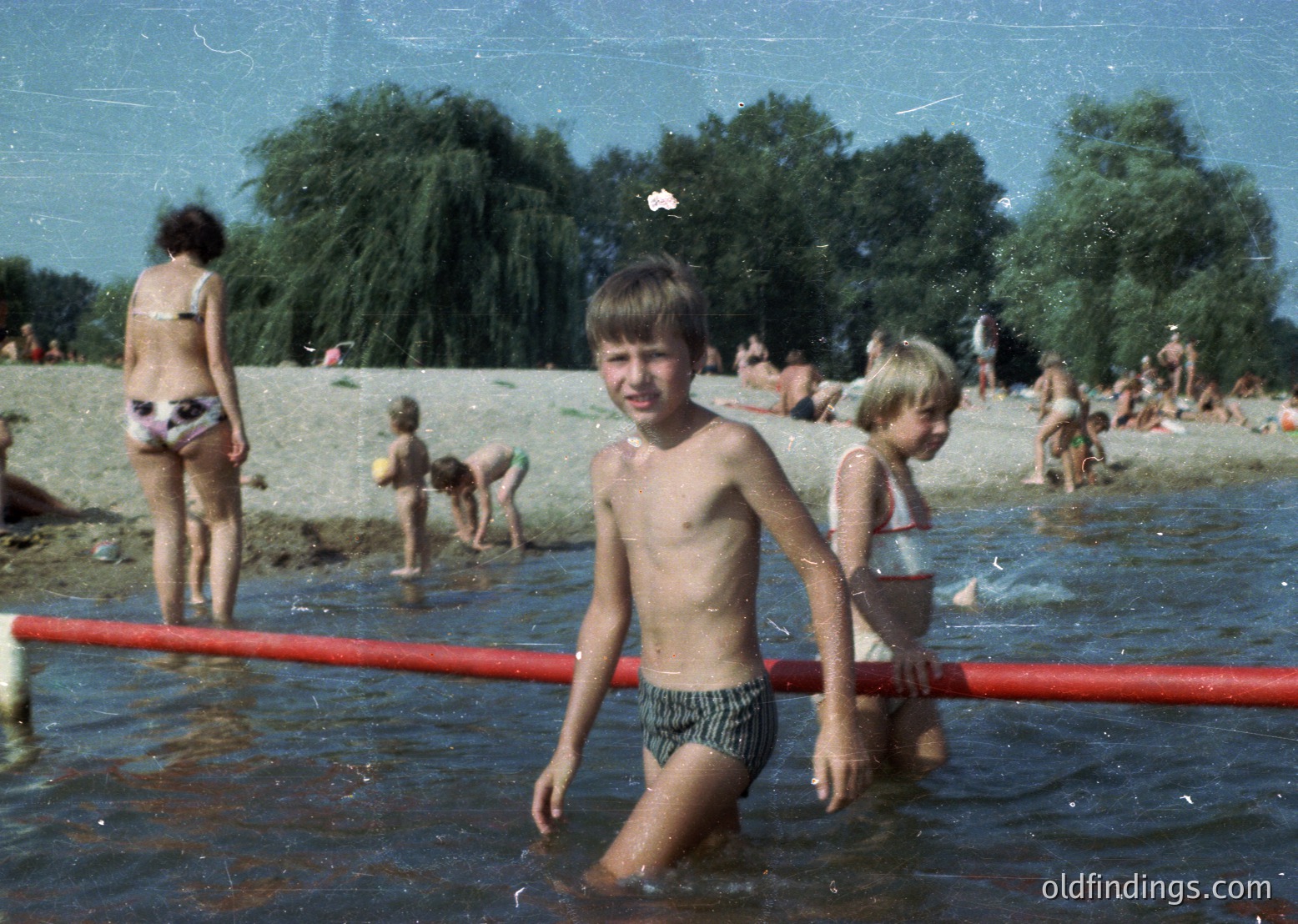 Vintage beach scene with children playing in shallow water, 1960s-1970s. Central boy in striped swim trunks stands near red pipe barrier, surrounded by sandy shore and lush greenery. Casual summer attire and relaxed postures reflect mid-century leisure culture.