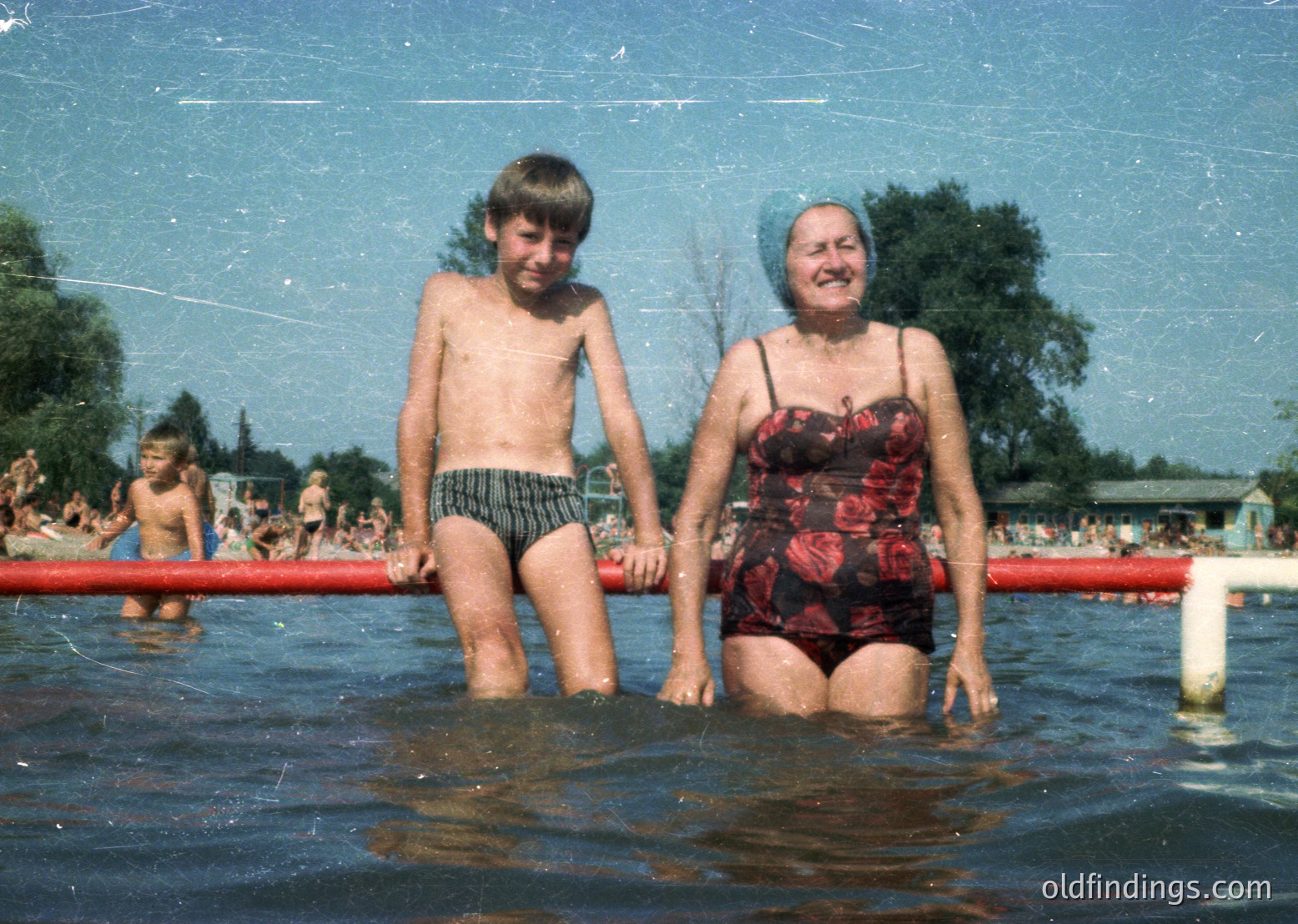 Vintage poolside scene featuring a woman in a floral swimsuit and headscarf holding a young boy in striped swim trunks. Crowded outdoor pool with red barriers and greenery in background, suggesting mid-20th century recreational setting.