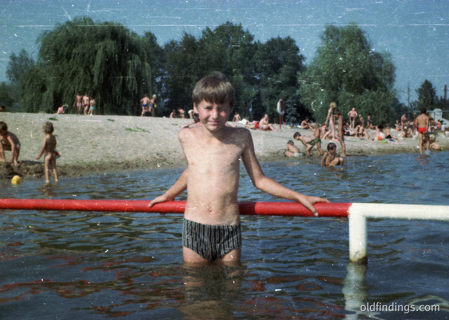 Shallow lakefront scene with a boy in striped swim trunks standing in water near a red pipe barrier. Lush green trees and grassy shore with other swimmers in background. Mid-20th century summer vibe.