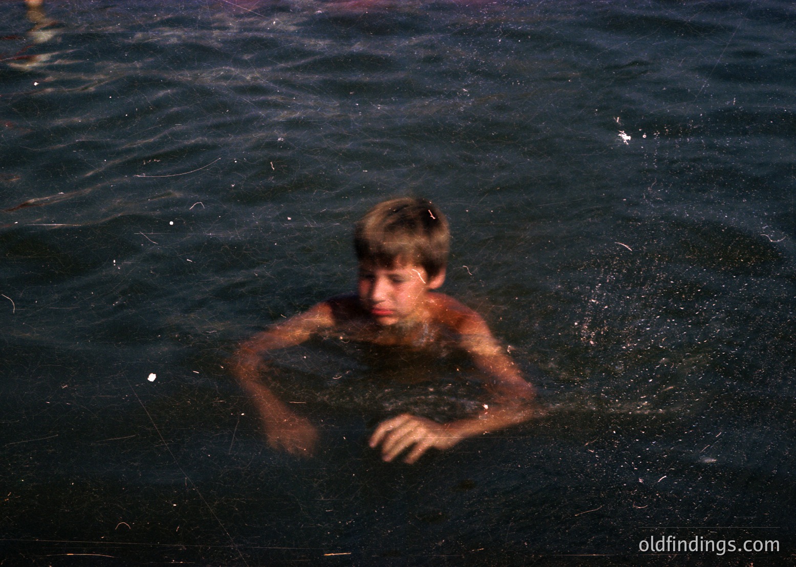 Young boy swimming in dark, shallow water, arms extended forward. Monochromatic, high-contrast tone suggests vintage or artistic filter. Likely coastal or lakeside setting.