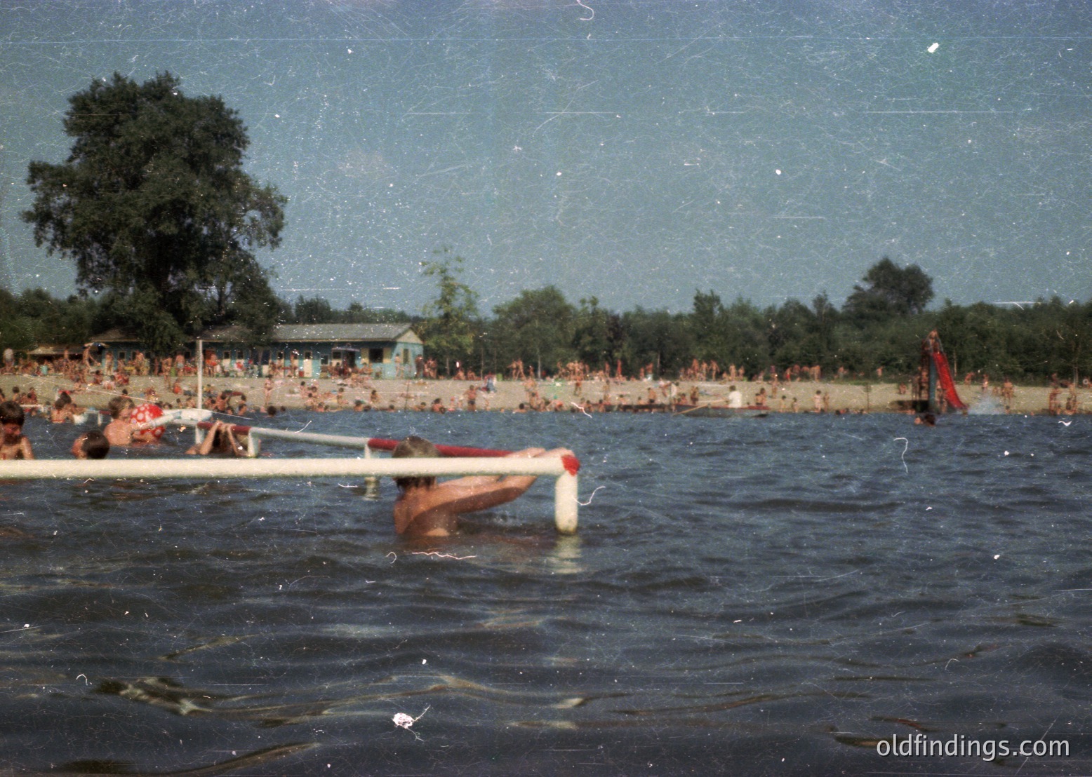 Vintage beach scene with crowded shore and waterfront. A lifeguard tower and wooden pier frame the foreground, while swimmers and sunbathers populate the sandy beach. Trees and a distant building suggest a lakeside or coastal resort. Film grain and faded colors indicate a mid-20th-century timeframe.
