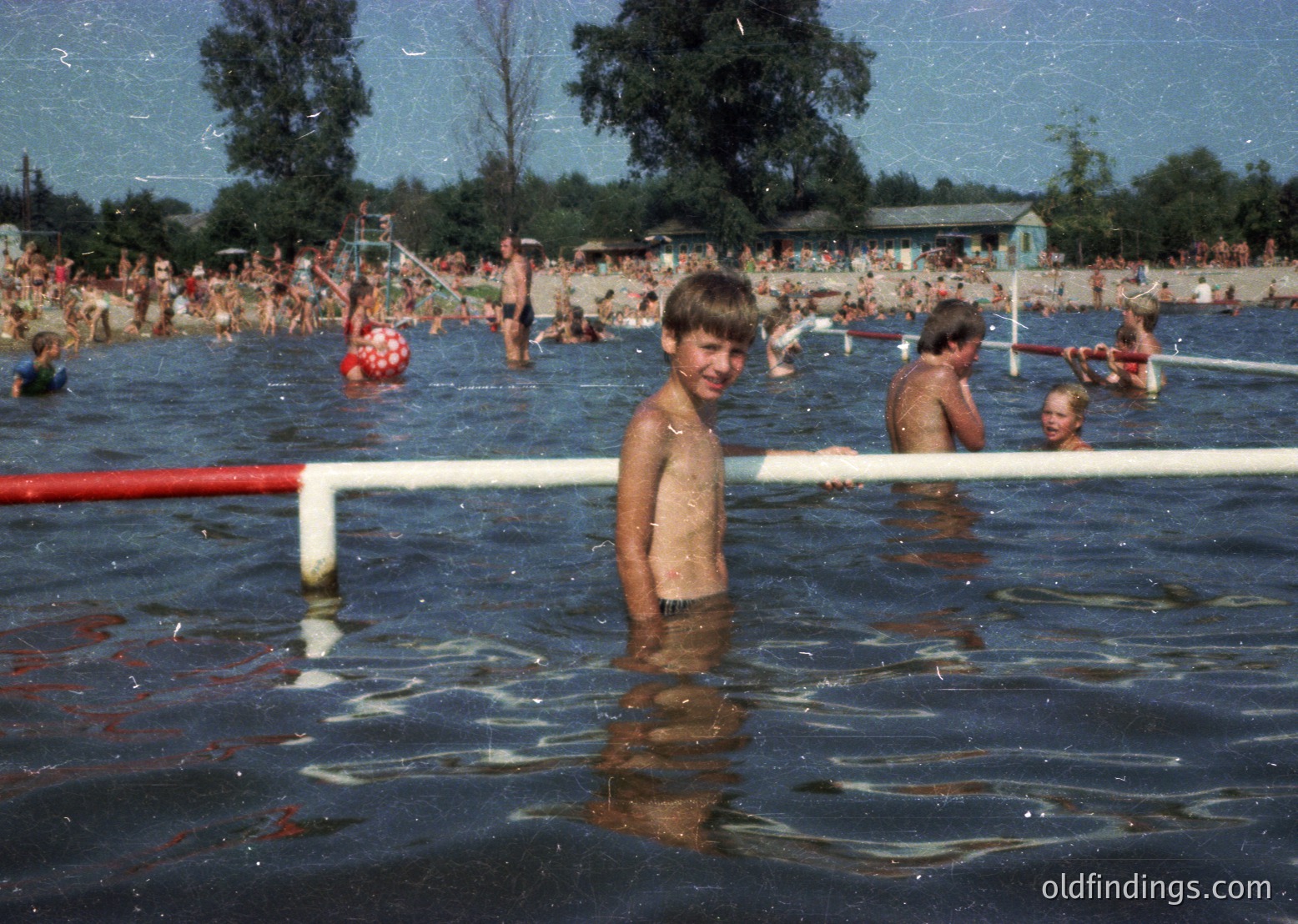 Vintage pool scene with young boy standing near a red-and-white goalpost in shallow water, surrounded by crowded swimmers. Lifeguard tower and trees in background suggest a public recreational area, likely mid-20th century.