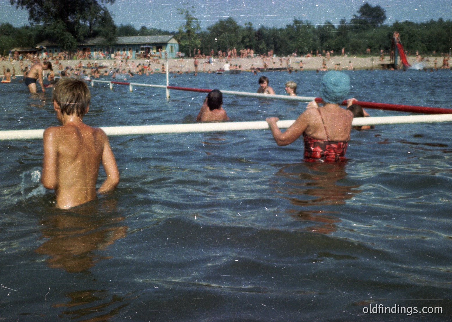 Vintage outdoor pool scene with lifeguard on duty, mid-20th century. Crowded beachfront with swimmers, wooden pool barrier, and red swim caps. Lifeguard in red trunks and blue cap supervises children playing in shallow water.