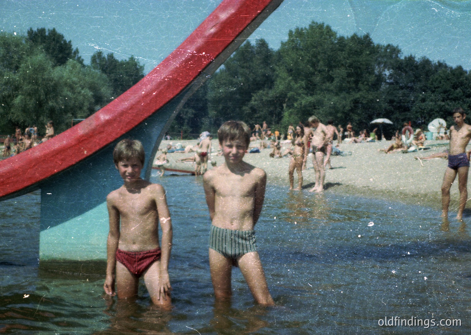 Two boys in wet swim trunks stand in shallow water under a red metal archway, likely part of a beach or pool entrance. Crowds of people relax on sand and in water in the background. Vintage sepia-toned photo suggests mid-20th century (1950s–1970s) recreational setting.