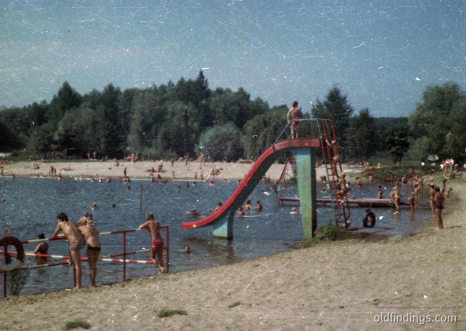 Vintage water park scene with red/blue slide and wooden platform, crowded with swimmers in 1960s-70s swimwear. Lush forested background suggests a lakeside resort.