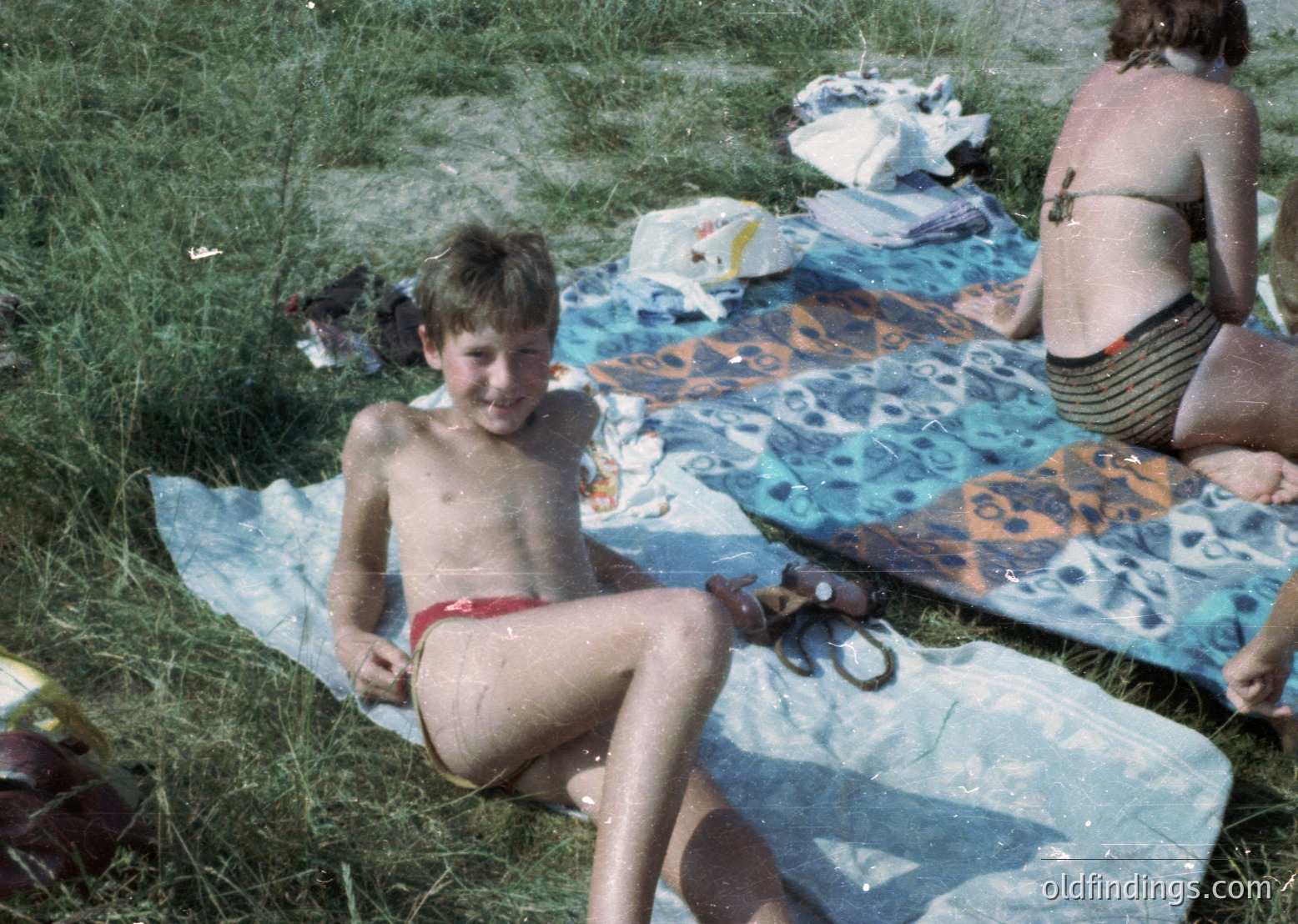 Vintage beach scene featuring a young boy in red swim trunks sitting on a patterned beach towel, surrounded by wet towels and scattered items. Likely 1970s European coastal setting.