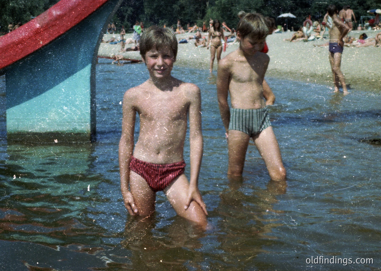 Two boys in red-striped swim trunks play in shallow water at a crowded beach, likely 1970s. Vibrant umbrellas and sunbathers fill the background, suggesting a public seaside resort. Authentic retro vibe captures mid-century beach culture.