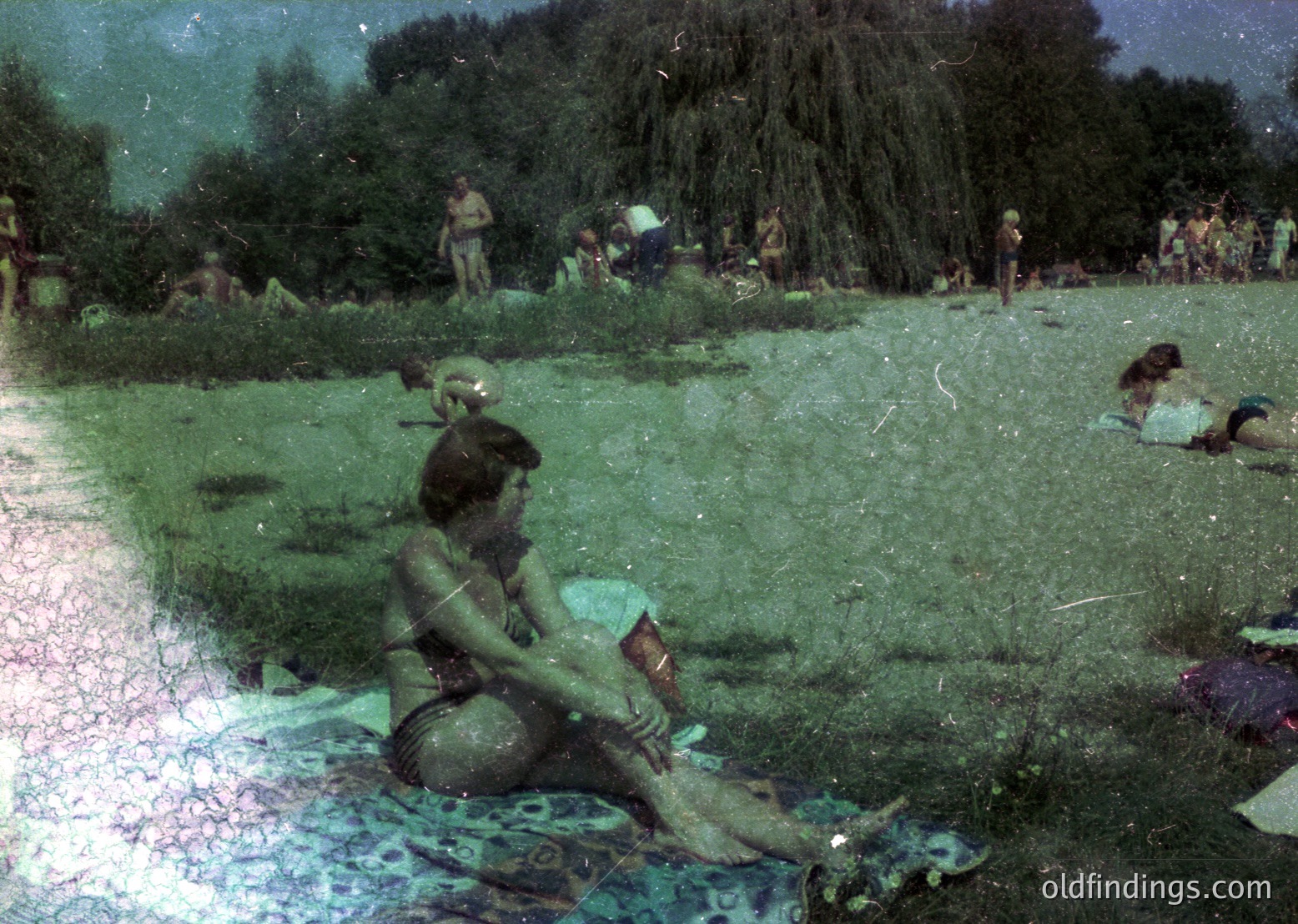 Vintage photo of a woman in a bikini reading a book on a patterned blanket in a grassy park. Mid-20th century summer scene with blurred crowd in background.