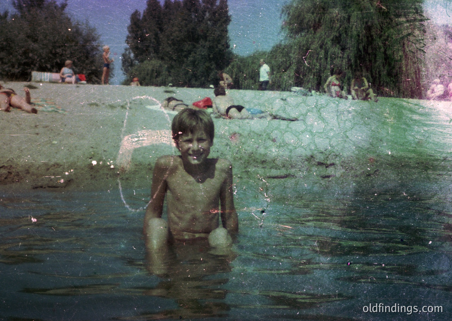 Vintage photo of a young boy playing in a shallow, algae-covered water body, likely a lake or river. Mid-20th century clothing (shorts) and greenscreen tint suggest or . Surrounding adults and trees indicate a communal outdoor setting.