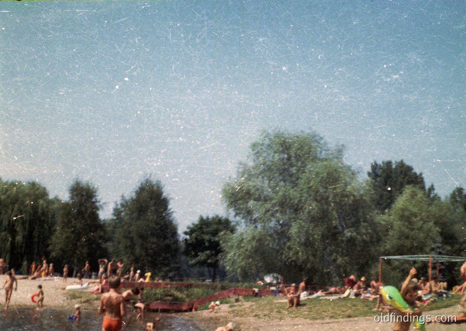 Vintage outdoor swimming scene with dense tree cover and clear skies. Mid-20th century attire—men in swim trunks, women in one-piece suits—suggests 1950s–1960s era. Wooden docks and scattered towels indicate a natural lake or riverfront. Warm, nostalgic aesthetic ideal for historical or retro-themed projects.