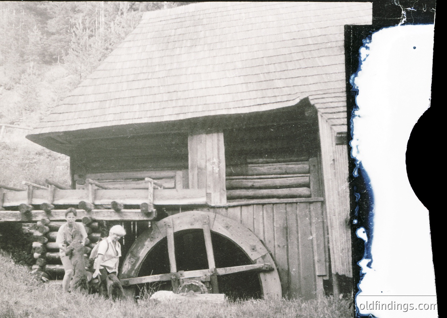 Vintage black-and-white photo of a rustic wooden waterwheel beside a log cabin, likely rural Europe, early-to-mid 20th century. Wheel’s wooden blades partially submerged in stream, with two men in period attire standing near. Log cabin features a sloped roof and wooden railings.