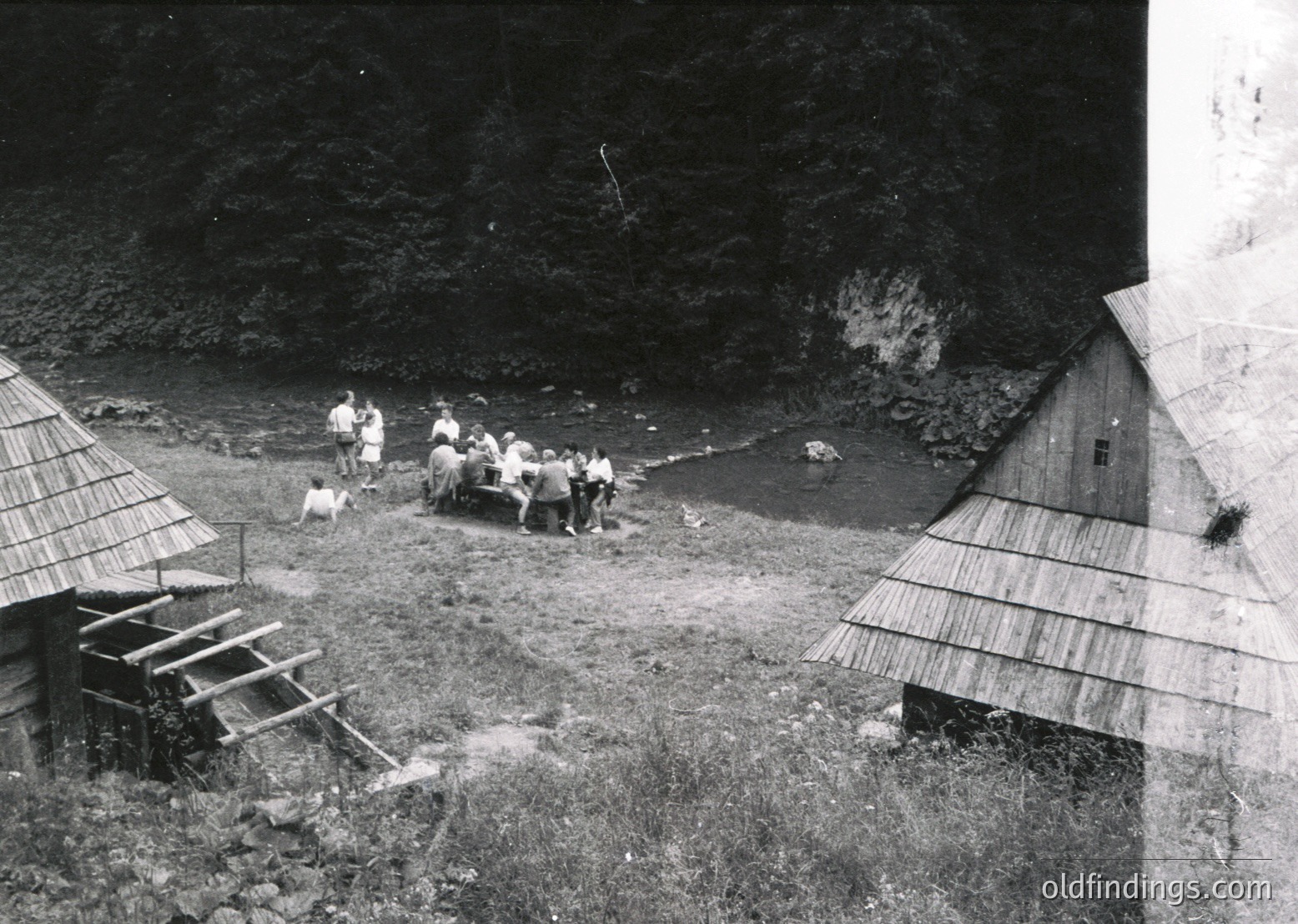 Black-and-white rural scene featuring a group of men in 1940s-50s attire gathering by a shallow stream, likely for water collection or communal work. Traditional thatched-roof huts with wooden planks flank the scene, set against a rocky hillside. Mid-20th century village life captured.