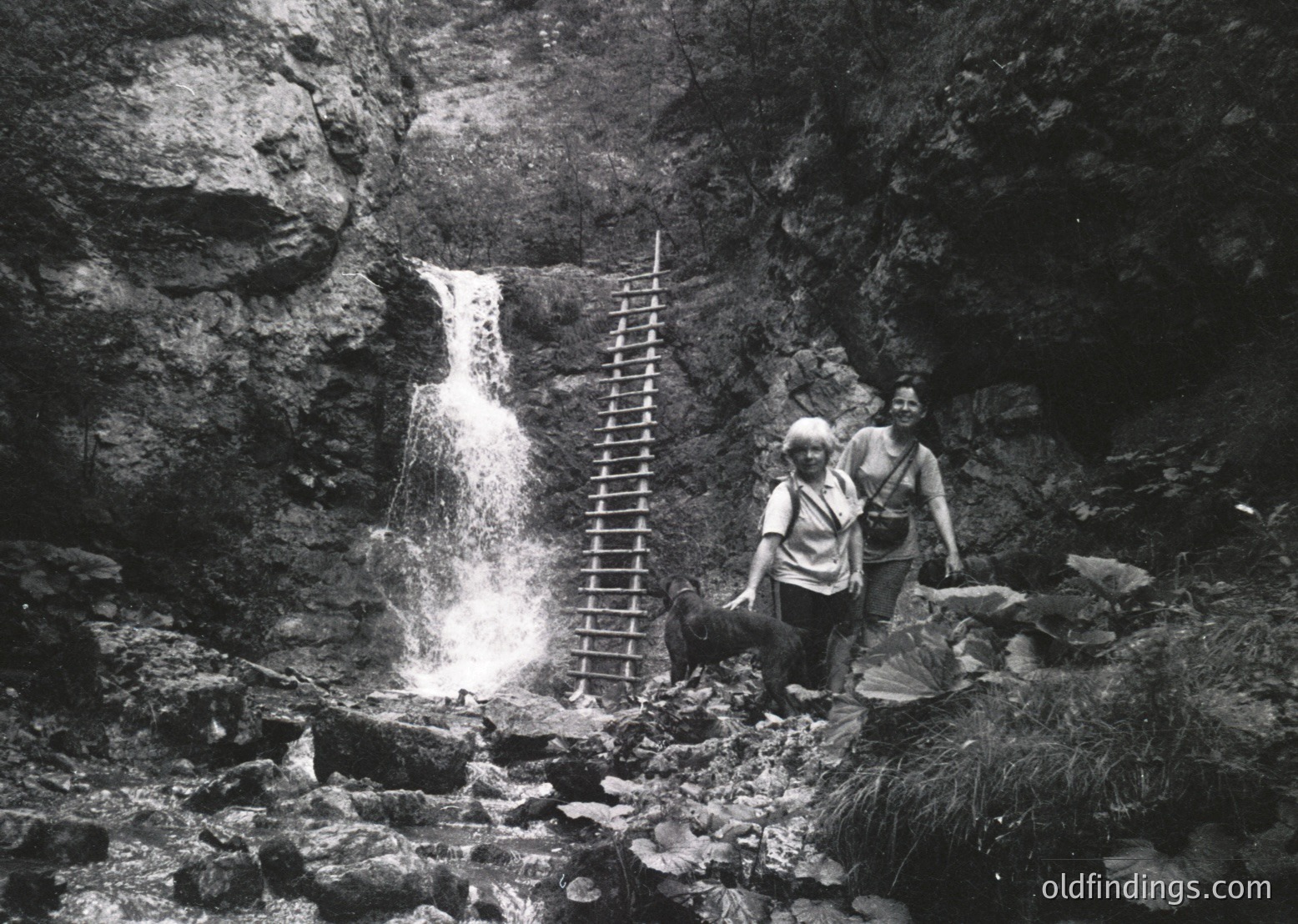 Mid-20th century black-and-white photo of two hikers beside a cascading waterfall in rugged alpine terrain. Ladder and rocky path lead to the falls, framed by steep rock walls. Hikers wear backpacks and wide-brimmed hats, suggesting outdoor adventure.