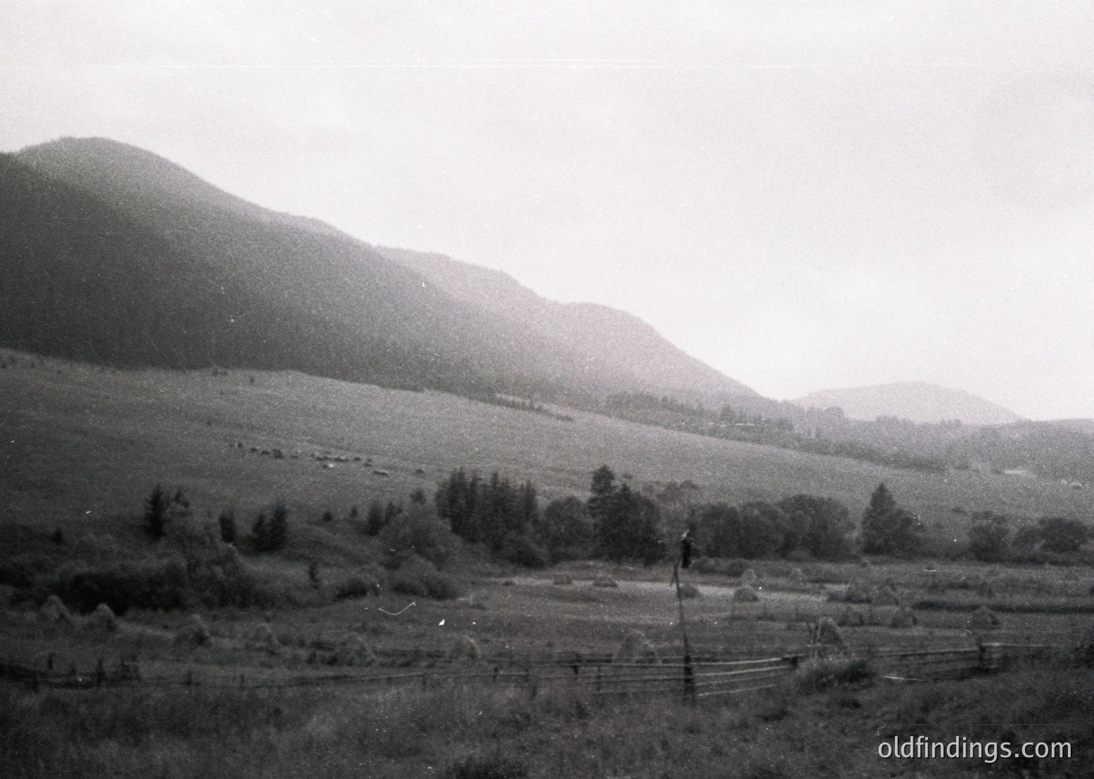 Vintage black-and-white rural landscape featuring rolling hills, sparse forest, and a single-track railway line winding through farmland. Low-lying structures and fenced fields suggest agricultural use. Foggy, atmospheric lighting enhances the mid-20th century feel.