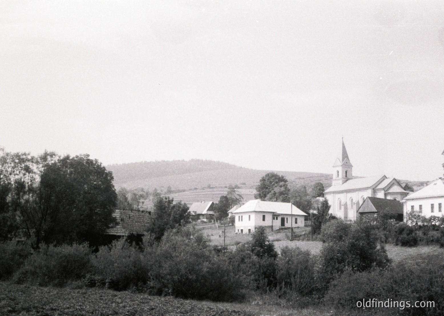 Black-and-white rural village scene featuring a prominent church with a tall steeple and whitewashed walls, surrounded by modest residential buildings. Rolling hills and dense greenery frame the horizon. Likely Eastern European, mid-20th century.