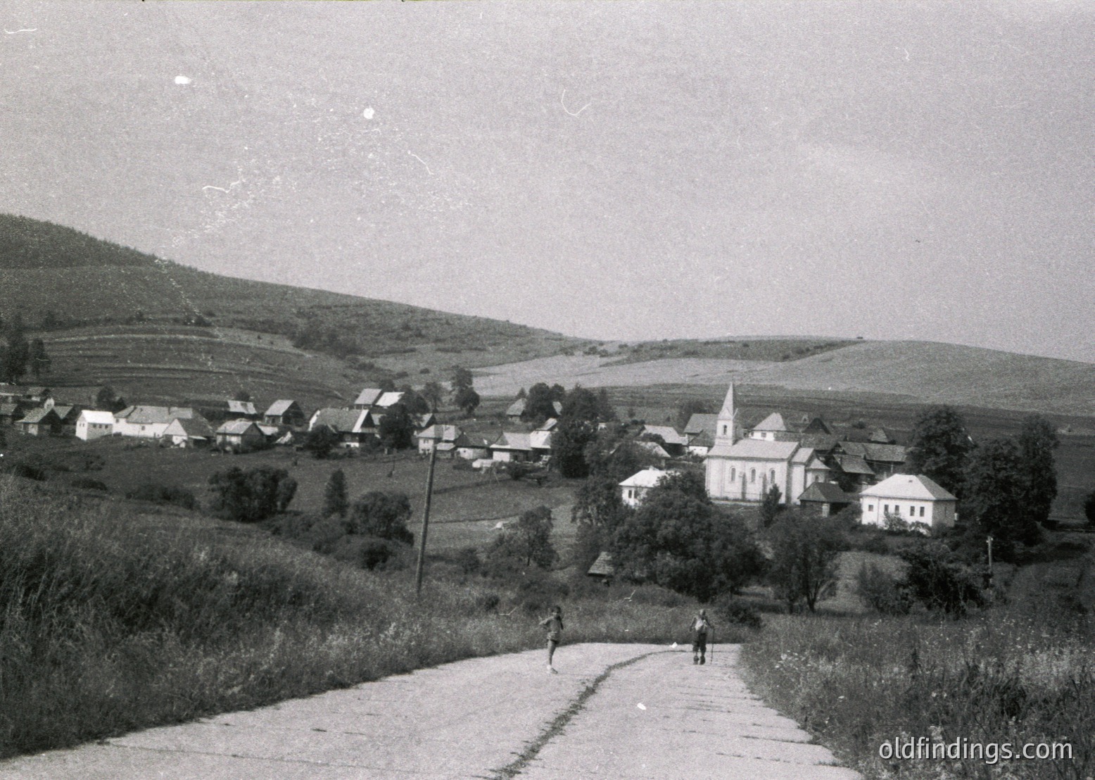 Vintage black-and-white rural village scene with a prominent church steeple and clustered houses on rolling hills. Two figures walking along a paved road, flanked by greenery and farmland. Likely Eastern European, mid-20th century.