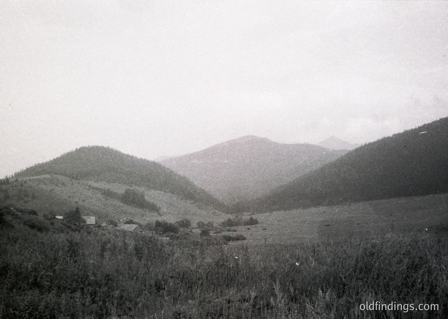 Vintage black-and-white landscape of rolling hills and sparse vegetation, likely European alpine terrain. Small, scattered structures suggest rural habitation, possibly agricultural. Fog or mist obscures distant peaks, enhancing dramatic atmosphere. Mid-20th century (1940s–1960s) style photography.