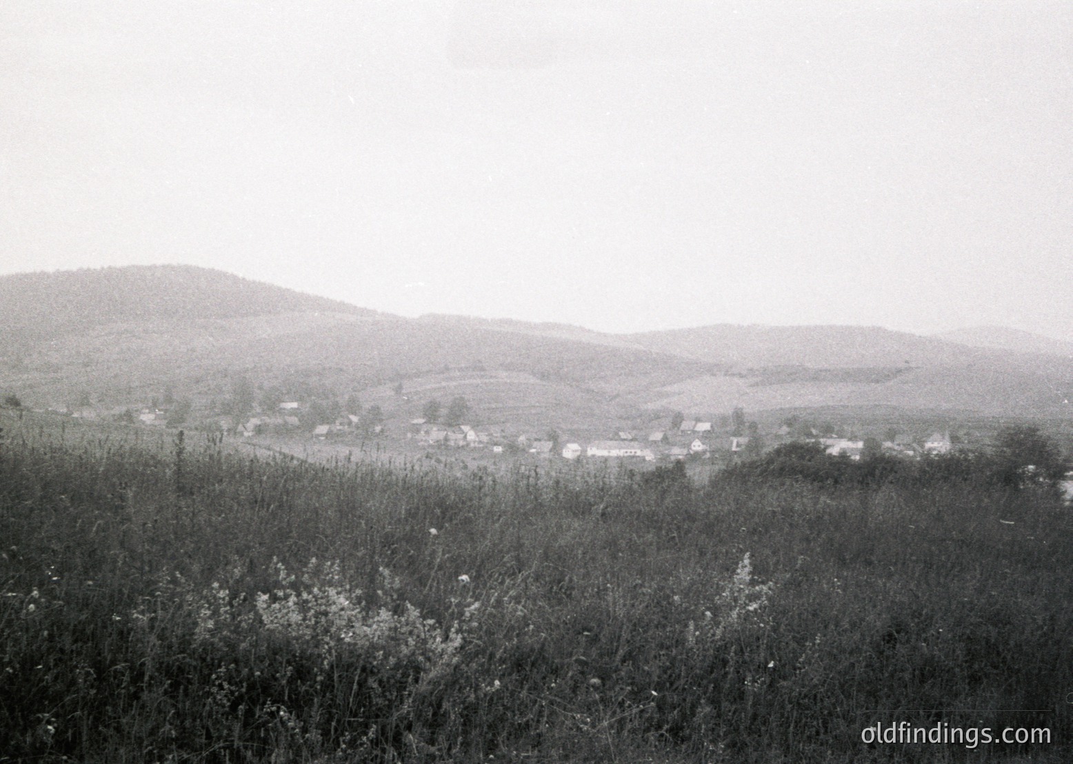 Mid-20th century rural landscape featuring scattered farmhouses and rolling hills. Low-contrast black-and-white composition highlights agricultural fields and sparse vegetation. Likely Eastern European countryside, 1950s–1960s.