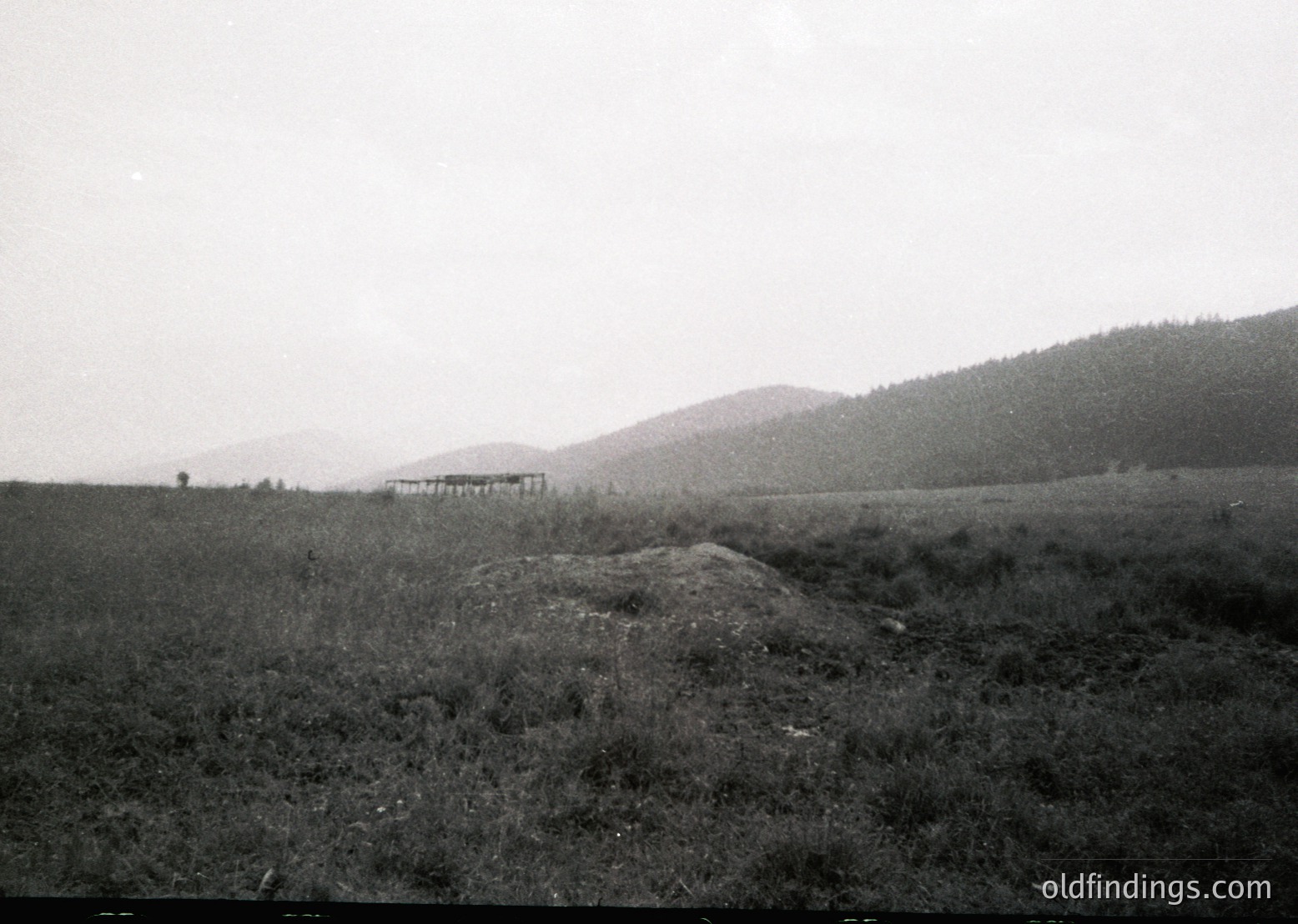 Vintage black-and-white rural landscape featuring a modest wooden structure on a slight hillside, surrounded by open fields and rolling hills. The hazy horizon suggests early 20th-century photography.