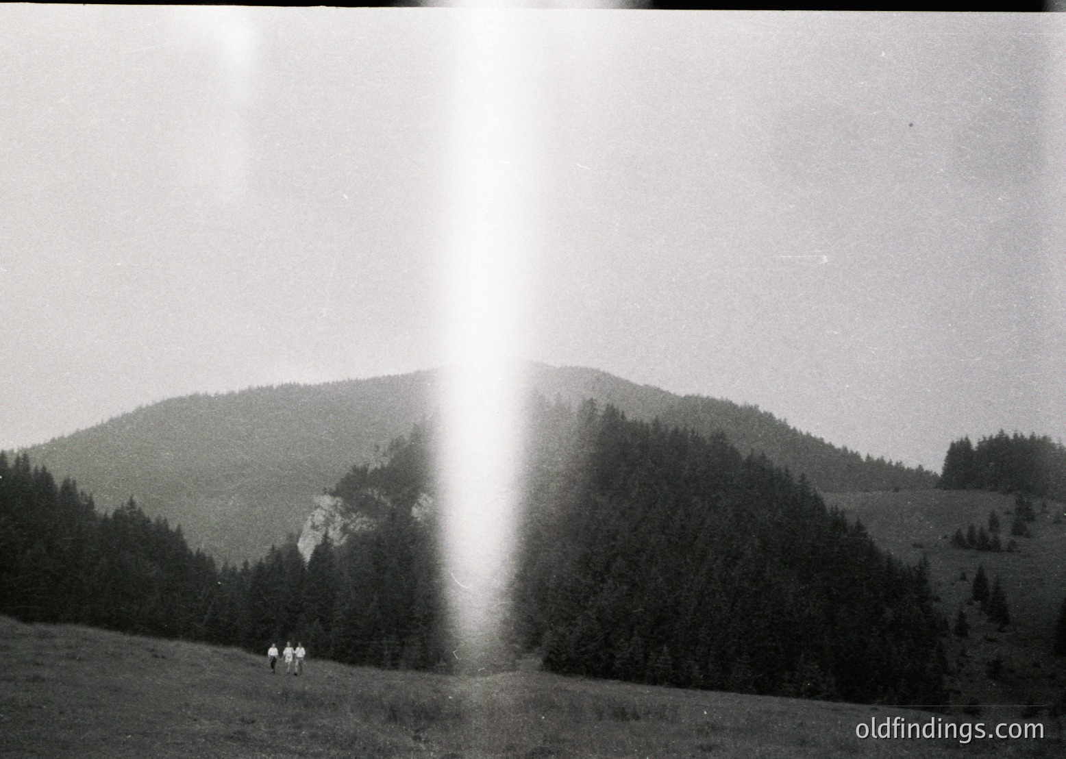 Vintage black-and-white mountain landscape with three hikers on a grassy ridge, framed by dense forest and rocky peaks. Sunlight creates a striking vertical light beam through the scene. Mid-20th century alpine hiking aesthetic.