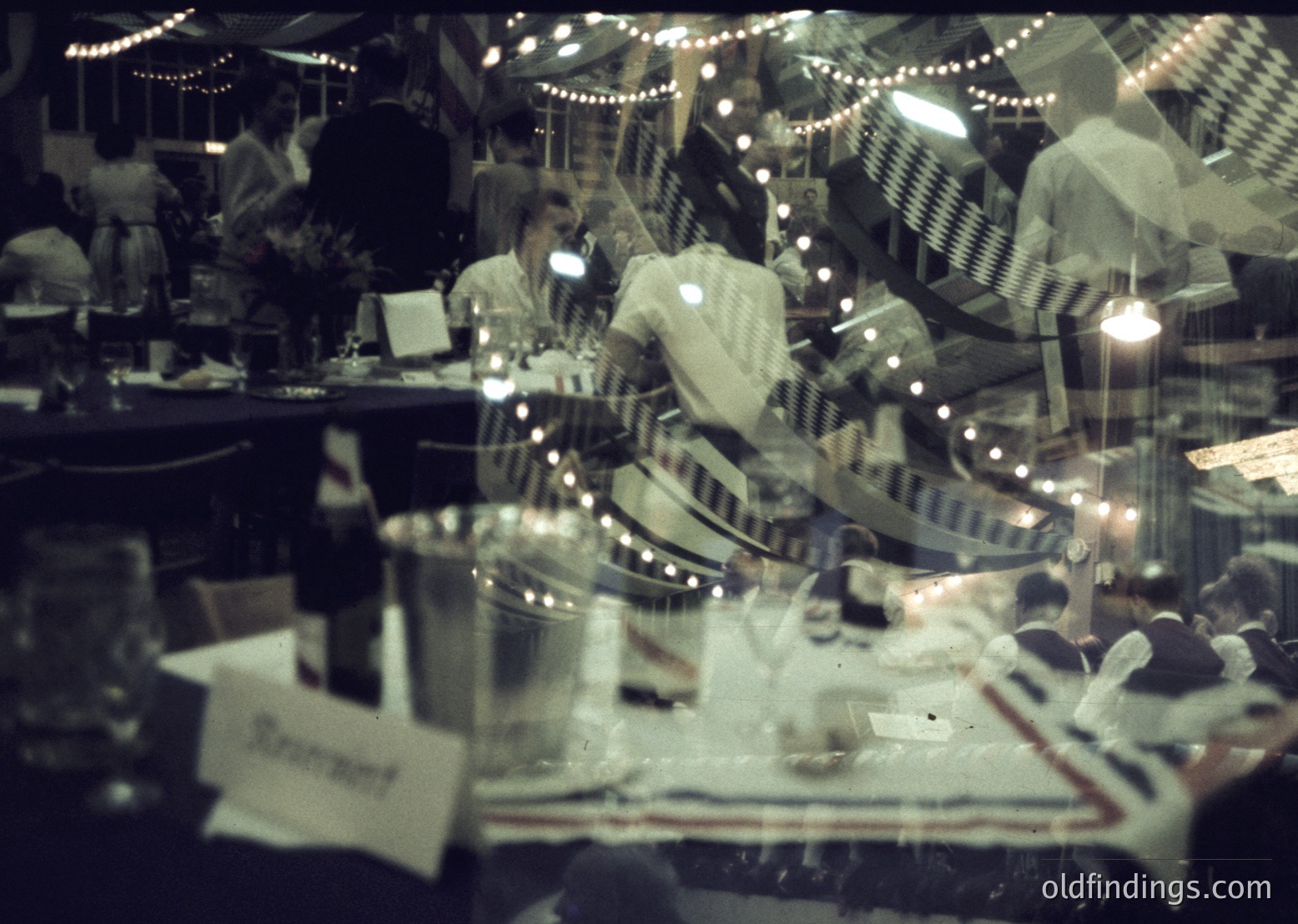 Vintage diner interior shot through a reflective surface, capturing blurred patrons in mid-20th-century attire. Neon signs, checkered tablecloths, and retro jukebox reflect in glass. Likely 1950s–1960s USA.