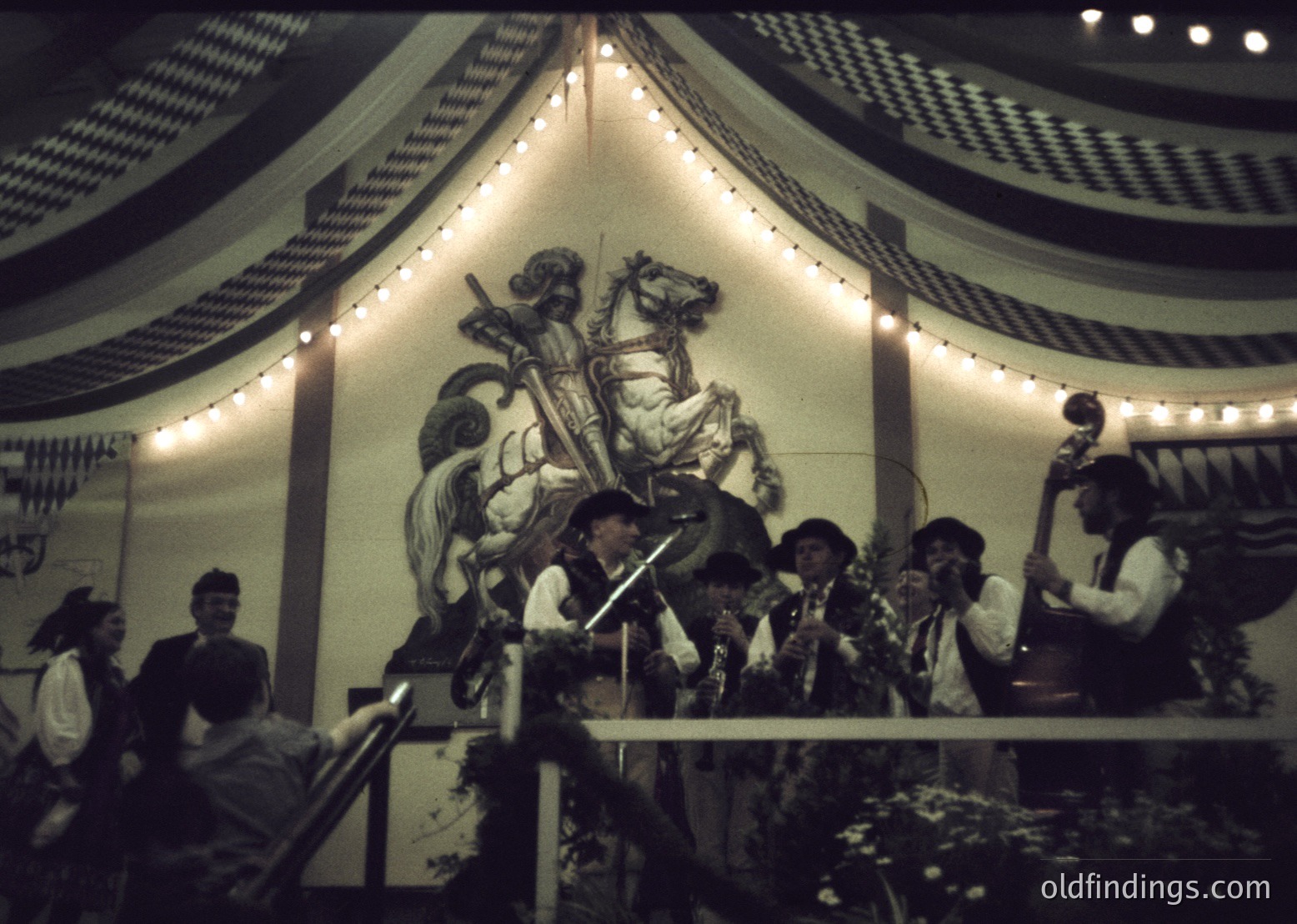 Vintage band performing under a decorative canopy with a mural of a knight on horseback. Stage adorned with string lights and floral arrangements. Audience seated in tiered seating, likely a 1960s–70s European cultural event.