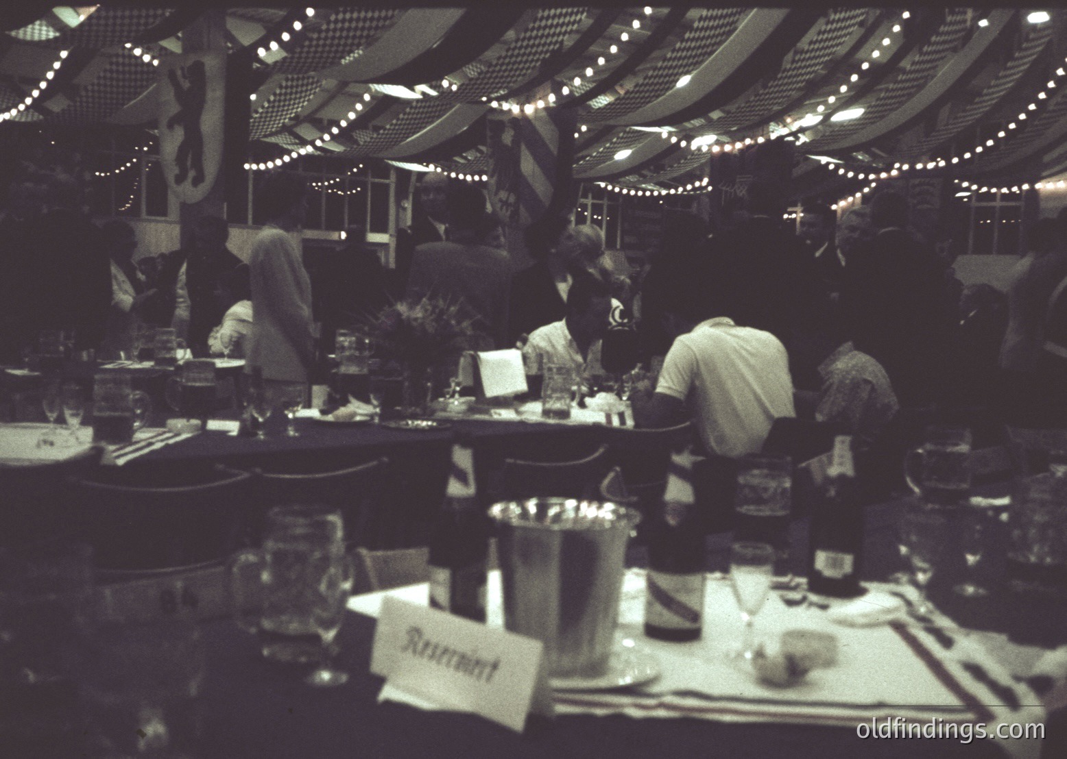 Indoor restaurant scene with string lights overhead, showcasing mid-20th-century dining culture. Round tables with place settings, wine glasses, and a "Restaurant" placard. Patrons in casual 1960s attire—men in suits, women in dresses—engaged in conversation. Warm, intimate ambiance with blurred motion suggesting lively atmosphere.