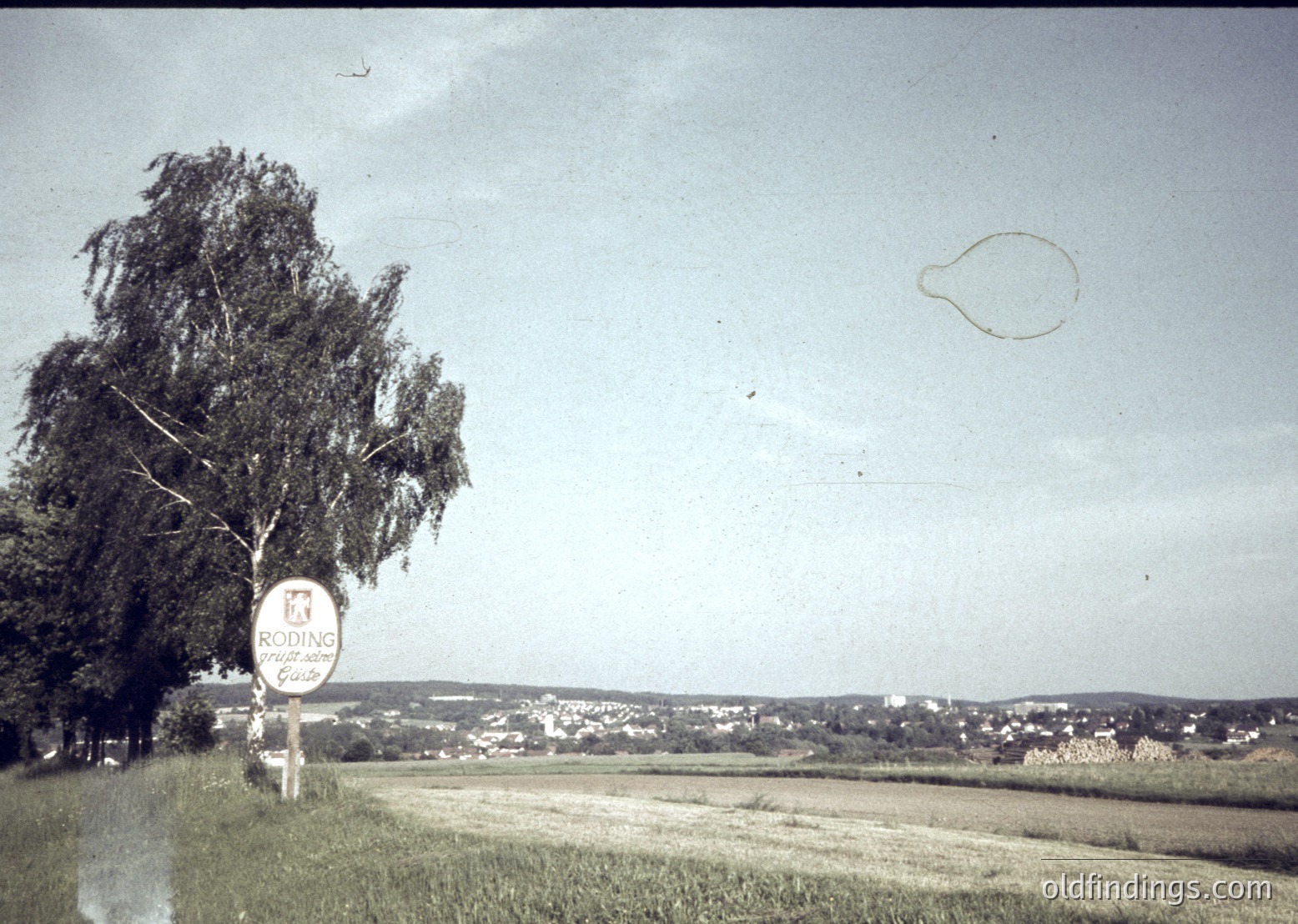 Vintage sepia-toned roadside sign reading **"CODINE"** (likely a brand) with a crown emblem, set against a flat, open landscape. Distant low-rise buildings and a lone tree frame the horizon. Likely Eastern European countryside, 1960s–1980s.