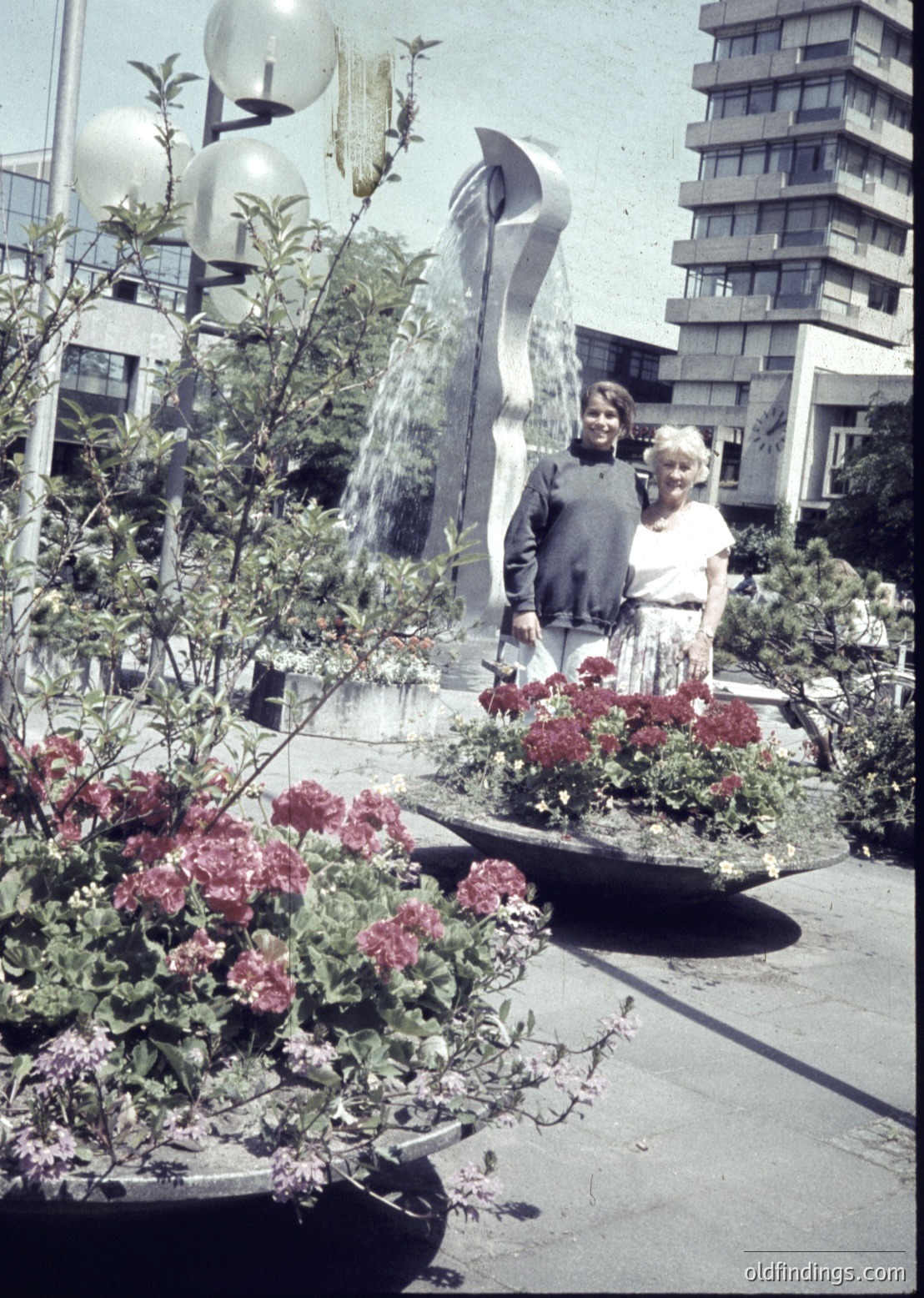 Two individuals pose beside a modernist fountain sculpture in a landscaped urban plaza, surrounded by vibrant pink geraniums. The background features Brutalist-style concrete buildings with spherical streetlights. Likely Eastern European, mid-20th century (1960s–1970s).