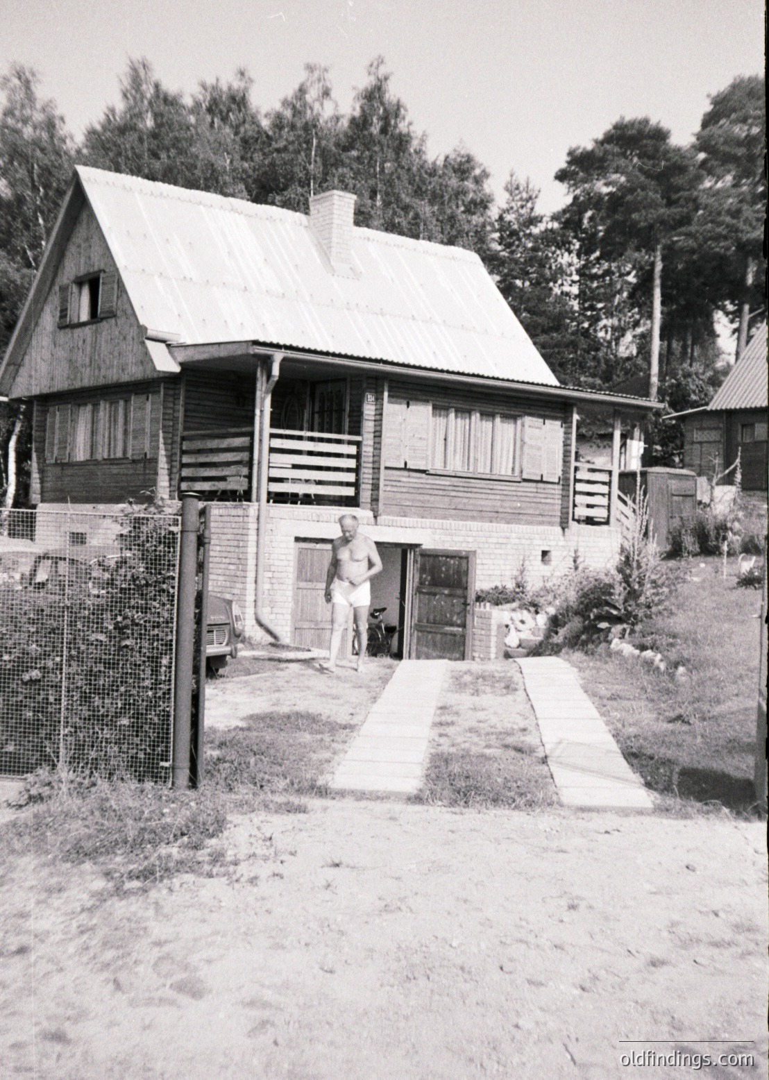 Mid-century alpine-style wooden chalet with gabled roof and brick chimney, set in a forested area. A man in shorts stands near the entrance, framed by a tiled pathway and wooden fence. Likely Eastern European, 1960s-70s.