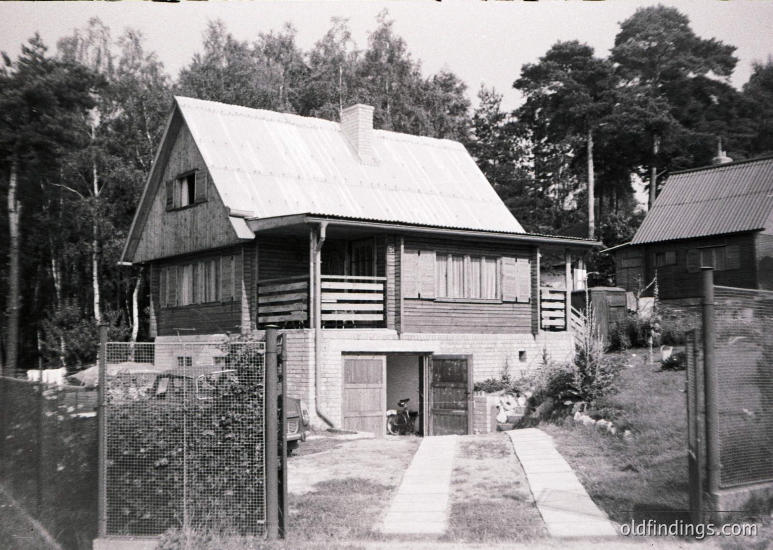 Mid-century alpine-style wooden cabin with gabled roof, set in a forested area. Log construction and brick chimney visible. Gravel driveway leads to a small garage/entryway with a bicycle parked inside. Fenced perimeter with chain-link and wooden posts. Likely 1950s–1970s European mountain retreat.