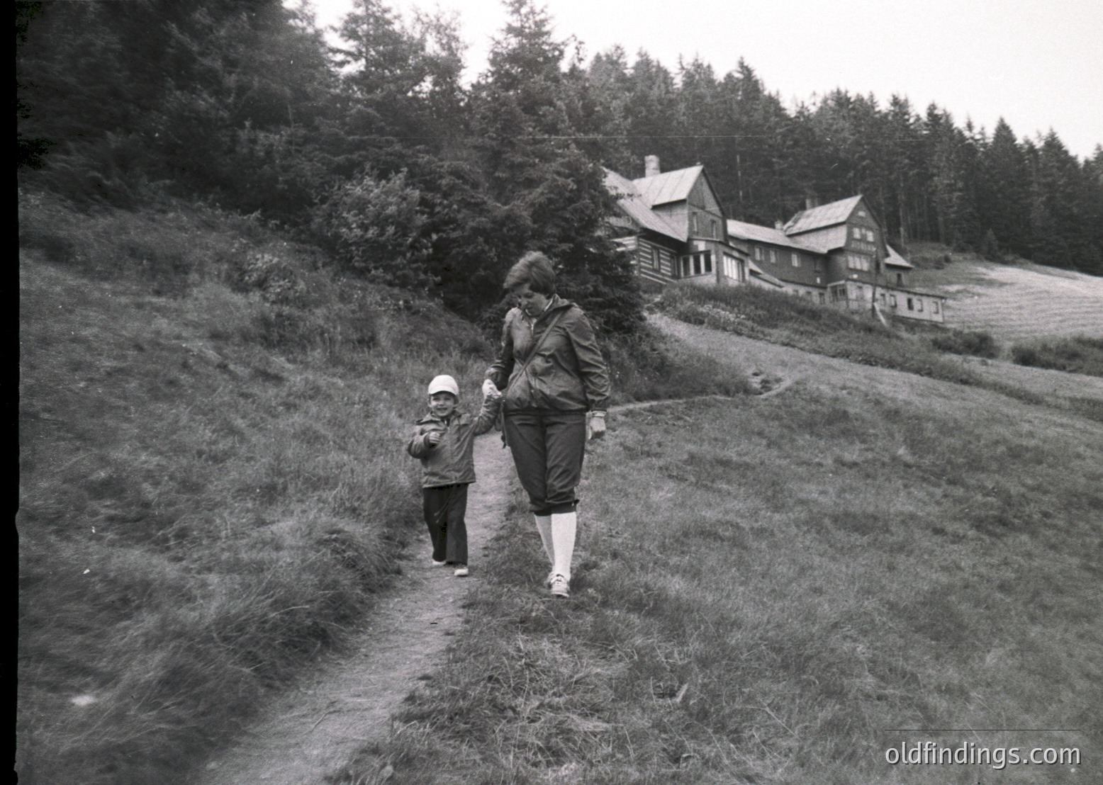Mid-20th century black-and-white photo of an adult and child walking on a grassy hillside path, leading toward rustic wooden chalets in a forested alpine setting. The adult wears a long coat and scarf, the child a hat and light jacket.