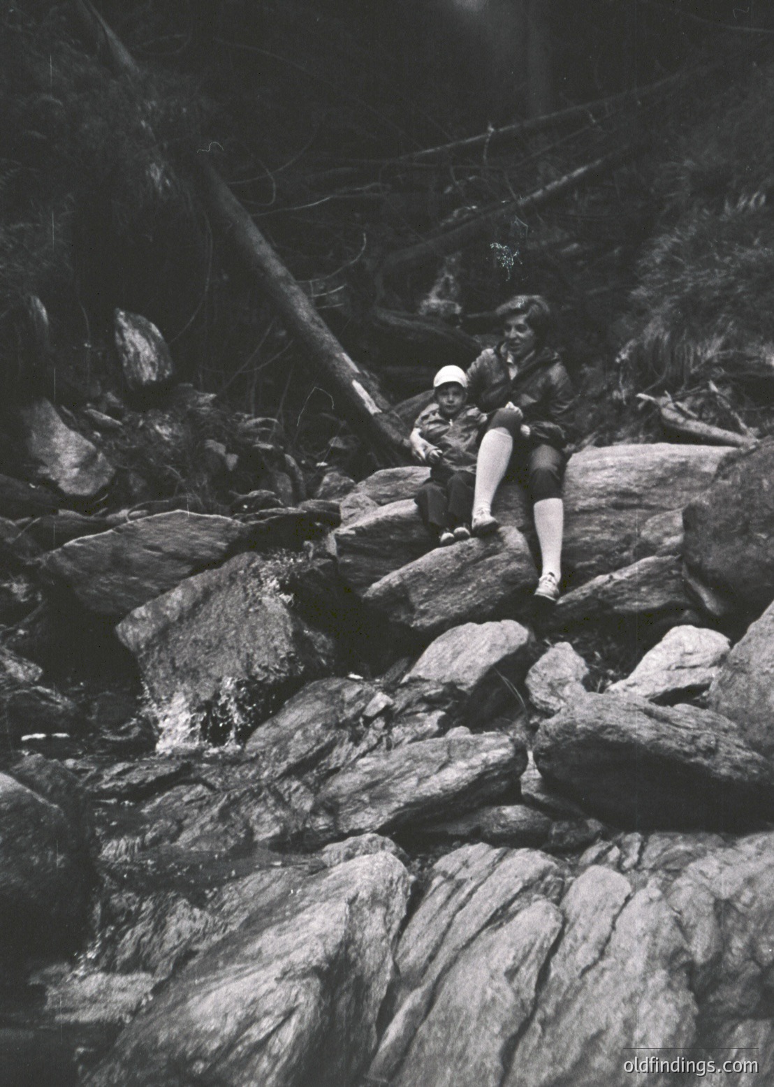 Mid-century outdoor portrait of a woman in a rocky canyon, seated on large boulders. She wears a wide-brimmed hat, short skirt, and knee-high socks, suggesting 1950s–1960s fashion. Sunlight filters through trees, creating dramatic shadows. Ideal for vintage travel or adventure photography.
