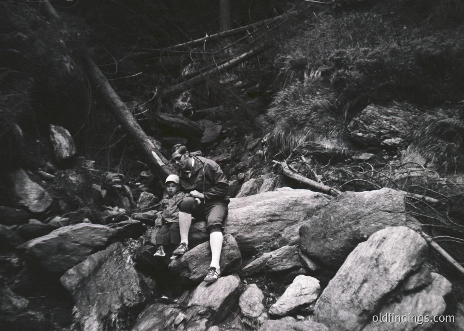 Black-and-white shot of a climber mid-ascent on a rugged rock face, secured by ropes and harness. The climber wears a cap, short-sleeve shirt, and hiking boots, gripping a rock with one hand. Suspended bridge and forest canopy visible in background.