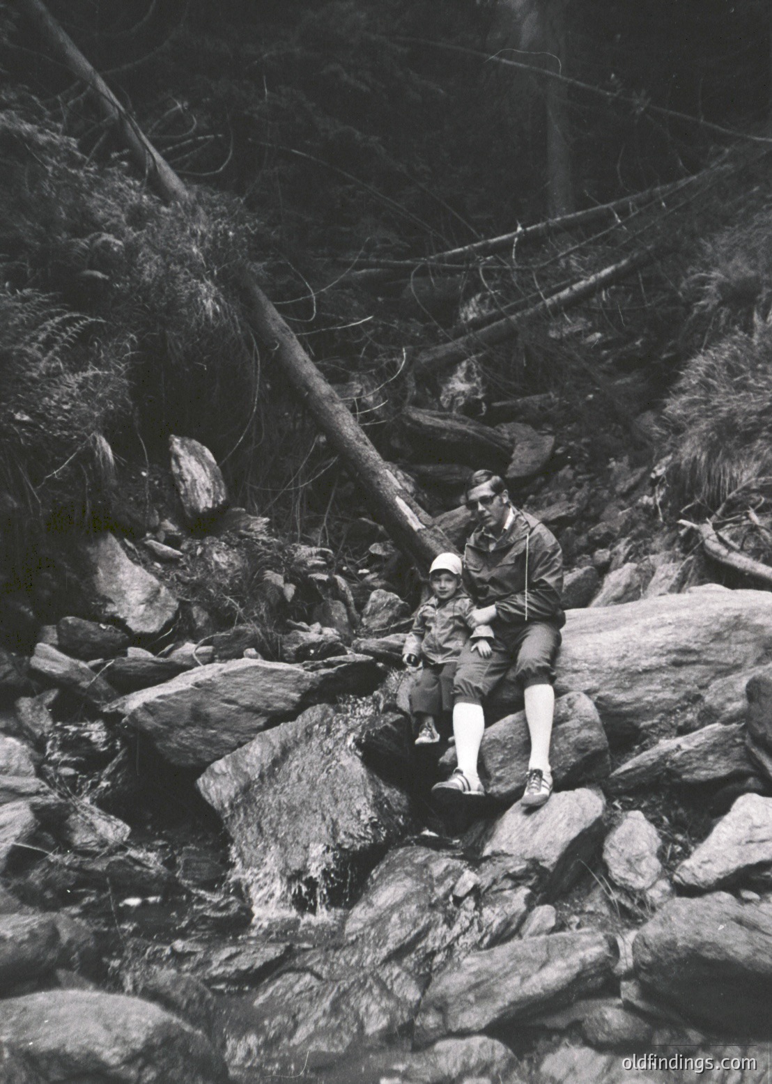 Two men in early 20th-century outdoor attire—wide-brimmed hats, long-sleeve shirts, and knee-high socks—rest on rugged rocks beside a shallow stream in a forested area. The man on the left holds a fishing rod, suggesting recreational angling. Dense foliage and fallen trees frame the scene, indicating a remote, natural setting. Likely mid-1900s, possibly or .