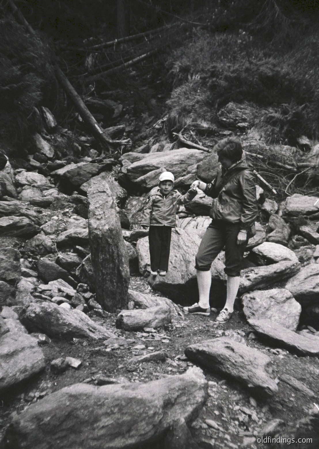 Mid-20th century black-and-white photo of two hikers navigating rocky alpine terrain. Adult assists child across uneven stones; both wear hiking gear (backpack, sturdy shoes). Dense forest and fallen logs frame the scene, suggesting a remote, natural setting. Style evokes mid-century outdoor adventure photography.