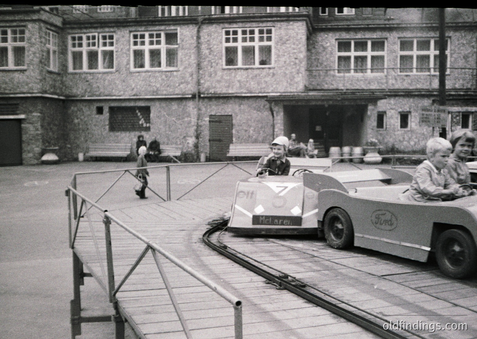 Vintage 1960s–70s courtyard scene featuring a Ford-based go-kart race with "McLaren" branding on a small kart. Spectators, including a woman in a white coat, watch from behind metal barriers. Brick building with large rectangular windows in background.