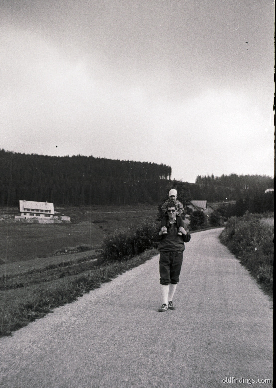 Mid-20th century rural roadside scene: Woman in 1950s-style dress and headscarf carries child on back, walking beside paved road flanked by fields and dense forest. Partial construction site visible in background.