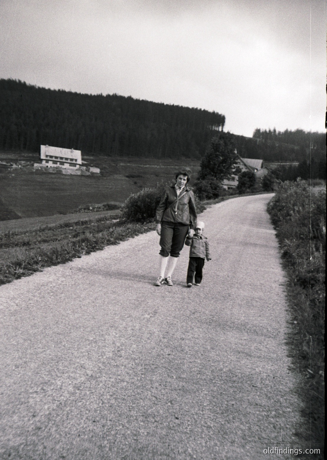A woman and child walk a rural road flanked by dense forest and open fields, likely mid-20th century. The adult wears a long coat, knee-high socks, and a headscarf; the child holds her hand. A distant building and farmland suggest agricultural life.