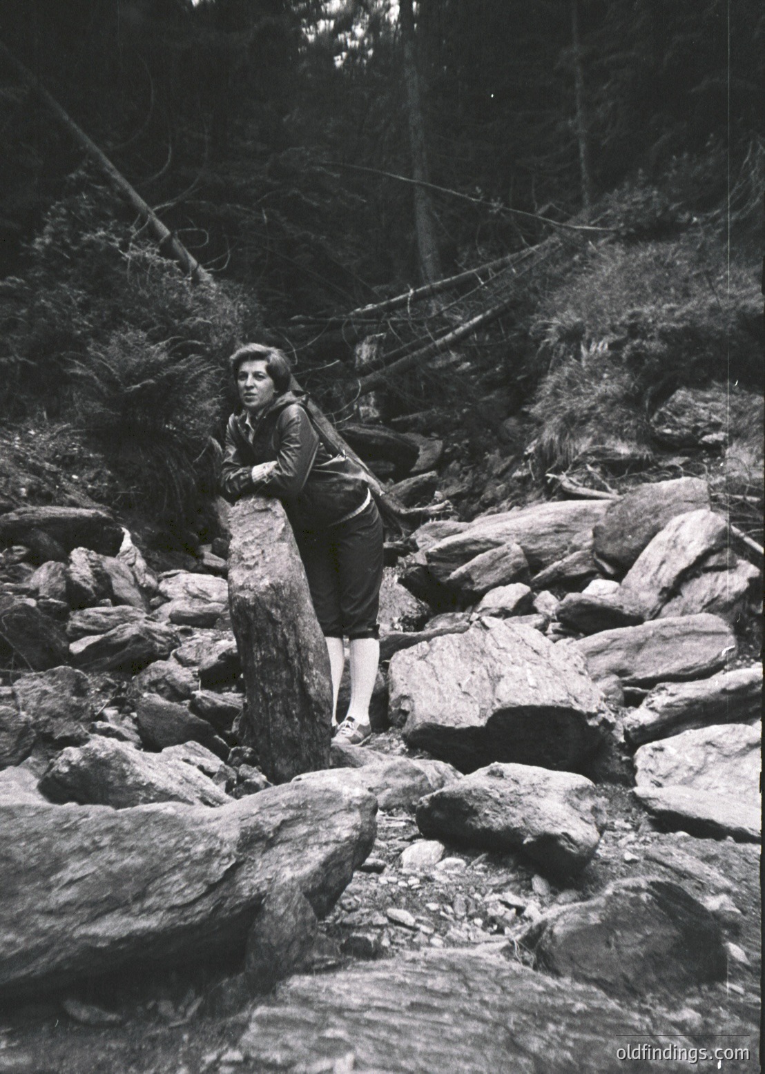 Mid-20th century black-and-white photo of a young woman in rugged forest terrain, gripping a fallen tree trunk for support. She wears a long-sleeve shirt, knee-length skirt, and knee-high socks. Surrounding rocks and dense pine trees suggest a hiking or exploration scene.