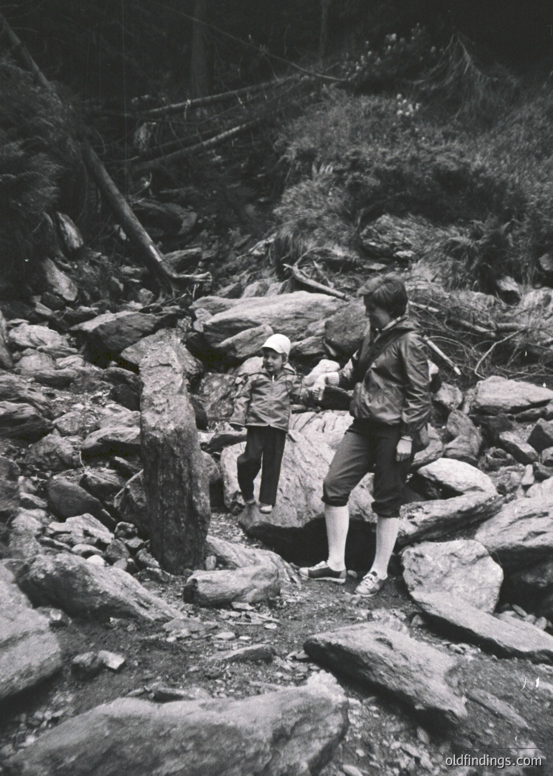 Two hikers navigate rocky terrain in a forested area, likely mid-20th century. The adult assists a child across a stream over flat stones. Clothing suggests outdoor adventure gear—knee-high socks, backpacks, and sturdy shoes.