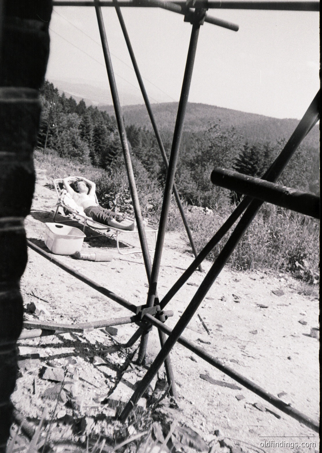 Mid-20th century alpine chairlift with rusted metal frame and cables, transporting a seated passenger through forested terrain. Snow-capped peaks and dense pine forests in background.