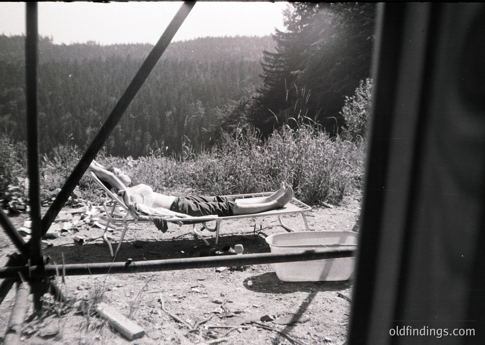 Mid-20th century black-and-white photo of a lone recliner positioned on a gravelly, forested clearing. A person lies relaxed in the chair, feet propped on a metal bucket. Dense pine forest and sunlit trees frame the scene, suggesting a remote, natural retreat.